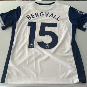 Tottenham Bergvall 15 matchtröja Nike - Tottenham Hotspur matchtröja med Bergvall 15 på ryggen. Se defekt på sponsorn på sista bilden. Vit med mörkblå detaljer och röda AIA-trycket på bröstet. Premier League-logga på ärmen, klubbmärke och Nike-logga framtill. Tillverkad i lätt, ventilerande polyester. Perfekt för dig som älskar Spurs. Endast handtvätt gäller. Replika
