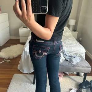 True Religion Becca Bootcut Jeans - Säljer ett par mörkblå True Religion Becca jeans med bootcut och låg midja. Jeansen har coola rosa kontrastsömmar och detaljerade bakfickor med lock och knappar. Det är knappt använda. Köpta för 2000kr. Pris kan diskuteras!