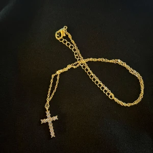 Korshalsband i guldfärg - Fint guldfärgat korshalsband. Fina stenar på korset. Aldrig använt, helt nytt. Kommer i rosa smyckespåse.