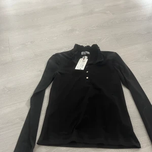 Svart långärmad topp från  Holly Whyte S - Svart långärmad topp från Holly Whyte by Lindex i mjuk bomull. Snygg volangkrage och tre guldfärgade knappar. Har aldrig använt. Därför prislappen är på🌸