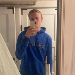 Blå Arigato hoodie  - Säljer en blå hoodie från Arigato med stort tryck över bröstet. Tröjan har huva med snörning och en klassisk känguruficka framtill. Perfekt för dig som gillar streetwear och vill ha en snygg och bekväm look.