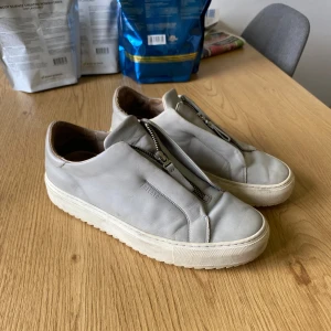 Grå sneakers från Axel Arigato med dragkedja - Snygga grå sneakers från Axel Arigato i skinn med unik dragkedja framtill istället för klassisk snörning. Skorna har en chunky vit platt sula och stilren design som passar till det mesta. Perfekta för dig som gillar minimalistisk och modern stil.