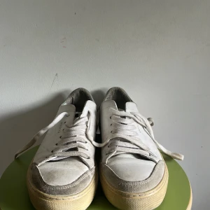 Vita sneakers från Axel Arigato - Snygga vita sneakers från Axel Arigato i storlek 42. Skorna har en klassisk låg siluett med snörning, beige sula och detaljer i mocka på tån. Tillverkade i skinn och mocka, med diskret guldtext på sidan och märkning bak.
