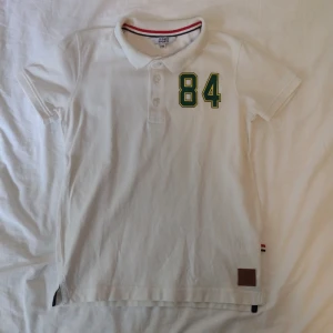 Vit pikétröja Iconic Sport med 84-tryck - Vit pikétröja från Iconic Sport med klassisk krage, tre knappar och grönt/gult 84-tryck på bröstet. Röd och marinblå rand på insidan av kragen och liten läderpatch nertill. Skön bomullskvalitet och korta ärmar, perfekt för en sportig look. Aldrig använd bara tvättad.