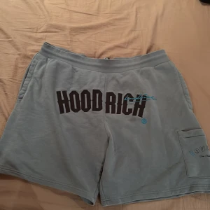 Blå Hoodrich shorts med tryck och T-shirt med tryck - Snygga ljusblå shorts från Hoodrich med stor svart logga framtill och blå detaljer. Mjukt material, elastisk midja och ficka på sidan med tryck. Perfekta för chill dagar eller streetwear-stil. Matchande T-shirt 