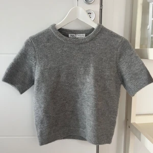 Grå stickad kortärmad tröja från Zara - Mysig grå stickad tröja från Zara med korta ärmar och rund halsringning. Tröjan har ribbade muddar vid ärmslut och nederkant, vilket ger en clean och enkel look. Perfekt att styla med jeans eller kjol för en chill vibe.
