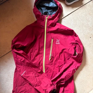 Röd vindjacka från Haglöfs  - Röd vindjacka från Haglöfs i storlek S. Jackan har huva, långa ärmar och en snygg kontrasterande gul dragkedja framtill samt på bröstfickan. Perfekt för blåsiga dagar och outdoor-äventyr. Lätt och smidig design med sportig känsla.