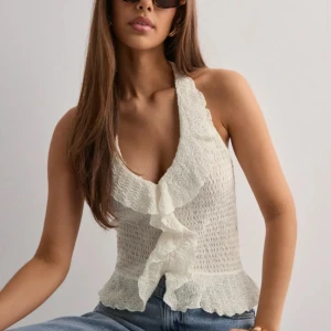 Nelly halterneck linnetopp med volang stl xs - Supersöt vit halterneck linnetopp med volangdetaljer framtill och vågig struktur. Toppen knyts i nacken och har en lätt croppad passform med volangkant nedtill. Oanvänd med prislapp kvar stl xs.