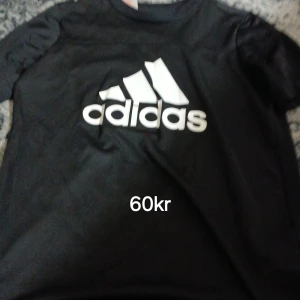 Svart Adidas t-shirt med logga - Svart t-shirt från Adidas med stor vit logga framtill. Klassisk rund hals och korta ärmar. Perfekt för dig som gillar sportig och enkel stil.