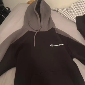 Champion hoodie i svart och grå - Säljer en snygg hoodie från Champion med svart kropp och grå ärmar och huva. Stor logga på ena ärmen och mindre logga på bröstet. Klassisk känguruficka och dragsko i huvan. Perfekt för chill eller streetwear.