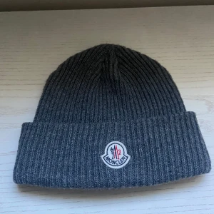 Moncler mössa - Ribbstickad grå mössa från Moncler med klassisk uppvikt kant och broderad logotyp framtill. Tillverkad i mjuk ull som håller dig varm och snygg under kyliga dagar. Perfekt accessoar för dig som gillar stilrena och exklusiva detaljer.