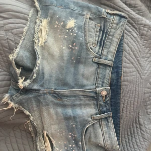 Blå jeansshorts med slitningar och pärlor - Snygga ljusblå jeansshorts med slitna detaljer och råa kanter. Shortsen har dekorativa pärlor och små blommor framtill, klassiska fickor och bälteshällor. Perfekta för sommaren och ger en cool, avslappnad vibe. PRIS GÅR ATT DISKUTERA‼️‼️