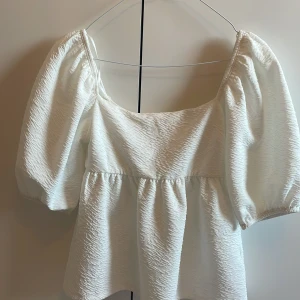 Vit blus med puffärmar och knyt - Supersöt vit blus med puffiga trekvartsärmar och struktur i tyget. Blusen har en fyrkantig ringning och knytning i ryggen för extra detalj. Perfekt för dig som gillar romantisk och trendig stil.