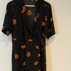 Svart omlottklänning med orange blommor - Svart omlottklänning från Ellos med kort ärm och knyt i midjan. Klänningen har ett coolt orange blommönster och v-ringad hals. Perfekt för dig som vill sticka ut med en färgklick och ändå hålla det stilrent.