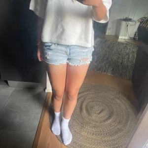 Ljusblå slitna jeansshorts - Säljer ett par ljusblå jeansshorts med råa, fransiga kanter och slitningar framtill. Klassisk femficksmodell med knapp och dragkedja. Perfekta för varma dagar och ger en avslappnad vibe. Snygga att matcha med en enkel t-shirt eller hoodie.