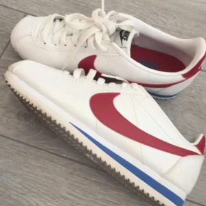 Nike Cortez  - Nike Cortez sneakers i vitt skinn med röd swoosh och blå detalj på sulan. Äkta samt helt oanvända. Ny pris 2000kr, pris går att diskutera vid snabbaffär!