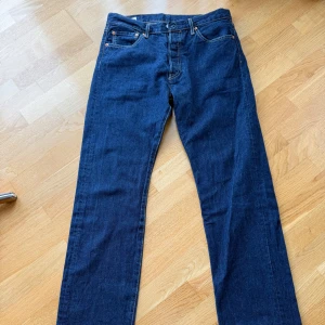 Levi's 501 mörkblå jeans W33 L34 - Klassiska Levi's 501 jeans i mörkblå denim med raka ben och femficksmodell. Snygga kontrastsömmar och ikonisk läderpatch bak i midjan. Perfekta för dig som gillar tidlös stil och vill ha ett par jeans som funkar till allt. ALDRIG ANVÄNDA.