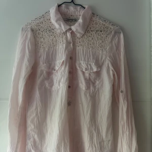 Ljusrosa skjorta med spets från Copenhagen Luxe - Supersöt ljusrosa skjorta från Copenhagen Luxe med spetsdetaljer över axlar och bröst. Skjortan har två bröstfickor med knappar, klassisk krage och långa ärmar. Perfekt för dig som gillar romantiska och feminina plagg.Titta på sista bikden, lite skavanker.