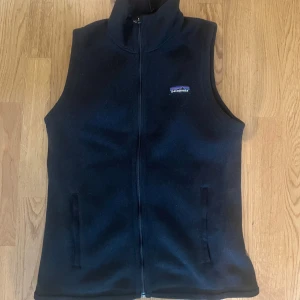 Svart fleeceväst från Patagonia - Svart fleeceväst från Patagonia i storlek S. Västen har hög krage, hel dragkedja framtill och två fickor. Tillverkad i 100% återvunnen polyester med mjuk, teddy-liknande insida. Perfekt för lager-på-lager och outdoor-vibe. Köpte den igår o säljer för att den ej passade 