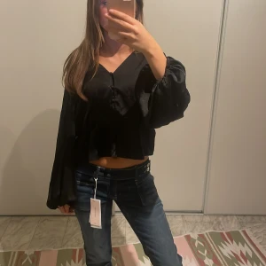 Svart croppad blus med vida ärmar - Säljer en svart croppad blus i glansigt material med v-ringning och knappar framtill. 