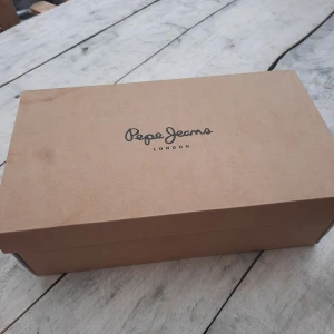 Mörkgrå sneakers från Pepe Jeans - Snygga mörkgrå sneakers från Pepe Jeans med ovandel i mocka och textil. Skorna har rund tå, platt sula och klassisk snörning. Perfekta för dig som gillar stilrena och sportiga skor med diskret logga på sidan. Som nya förutom undersidan på hö sko som ni ser på sista bilden. Har blivit en liten grop/avskavning i sulan, inget som påverkar.