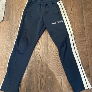 Palm Angels trackpants med vita ränder - Mörkblå trackpants från Palm Angels med vita dubbla sidoremsor och logga på låret. Säljer på grund av de inte är min stil, använt kanske 4-5 gånger så riktigt bra skick på dom😁
