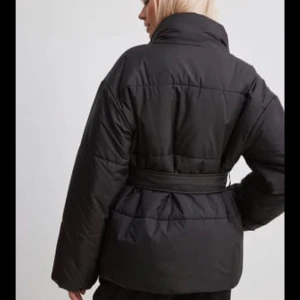 Svart pufferjacka med bälte - Säljer en svart pufferjacka med bälte i midjan och hög krage. Jackan har quiltad design, dragkedja och knappar framtill. Materialet är glansigt och känns riktigt mjukt och skönt. Perfekt för kalla dagar när du vill vara både varm och snygg.