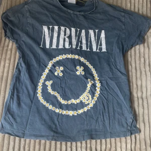 Blå Nirvana t-shirt med blommigt tryck - Snygg t-shirt med Nirvana-tryck i vitt och en stor smiley gjord av små blommor på framsidan. Klassisk rund hals och korta ärmar. Perfekt för dig som gillar bandtröjor och vill sticka ut med en cool, avslappnad stil.