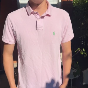 Ljusrosa pikétröja från Ralph Lauren - Säljer en ljusrosa pikétröja från Polo Ralph Lauren i bomull. Klassisk krage, korta ärmar och grönt broderat logotyp på bröstet. Custom fit-modell som sitter snyggt och passar perfekt till jeans eller shorts.