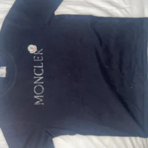 Svart Moncler t-shirt med logga - Svart t-shirt från Moncler med stor vit logga framtill och klassisk rund hals. T-shirten har korta ärmar och är tillverkad i mjuk bomull. Perfekt för dig som gillar stilrena och exklusiva plagg med tydlig branding. Den är lite liten i storleken ⚜️