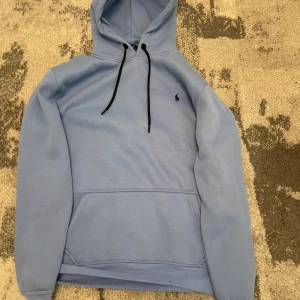 Ljusblå hoodie från Polo Ralph Lauren - Snygg ljusblå hoodie från Polo Ralph Lauren med svart broderad logga på bröstet. Tröjan har huva med svarta snören och en stor magficka. Perfekt för dig som gillar klassisk streetstyle och vill ha något bekvämt och stilrent. Pris kan diskuteras vid snabb affär