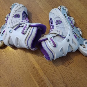 Vita och lila inlines med skydd - Säljer ett par snygga inlines från GRAF i vitt och lila med justerbara spännen och meshdetaljer. Medföljer svart hjälm och Tony Hawk skydd för knän, armbågar och handleder. Perfekt för dig som vill rulla fram med stil och säkerhet. Passar barn med storlek mellan 33-37.