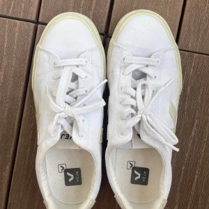 Vita Veja sneakers i canvas - Snygga vita Veja sneakers i canvas med klassisk V-logga på sidan och beige detaljer vid häl och sula. Skorna har rund tå, snörning och platt sula. Perfekta för en clean och stilren look.🩷