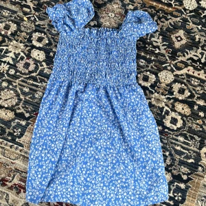 Blå blommig klänning - Charmig blå klänning med vita blommor och korta puffärmar. Den har en smockad överdel som ger en bekväm passform. Perfekt för sommarens varma dagar!