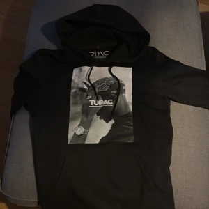 Svart hoodie med Tupac-tryck från Mister Tee - Säljer en svart hoodie från Mister Tee med ett stort Tupac-tryck på framsidan. Hoodien har en klassisk passform med långa ärmar och en bekväm huva. Perfekt för dig som gillar streetwear och vill ha en stilren look.