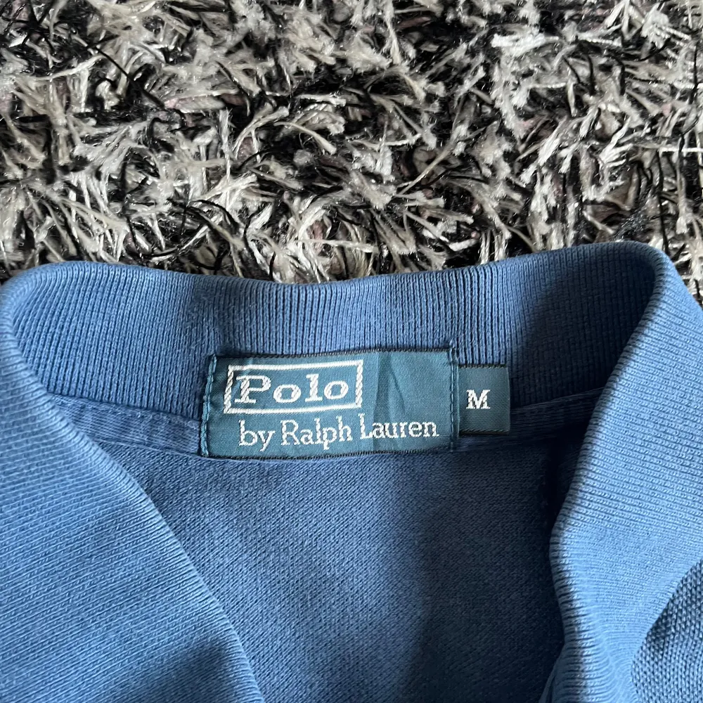 Säljer en klassisk blå pikétröja från Ralph Lauren med den ikoniska loggan broderad i gult på bröstet. Tröjan har korta ärmar och en stilren krage med knappar. Perfekt för en avslappnad och stilren look. Skick 9/10. Nypris runt 1500kr. T-paidat.