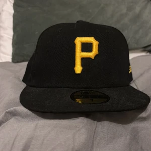 Svart keps från New Era - Snygg svart keps från New Era med ett broderat gult 'P' framtill. Kepsen är en 59FIFTY-modell och har MLB-loggan broderad baktill. Perfekt för sportiga tillfällen eller som en stilren accessoar. Nyskick, aldrig använd. Storlek 7 - 55,8cm. 