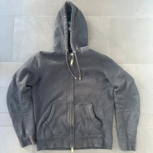 Burberry Hoodie - Säljer denna snygga mörkgrå Burberry zip hoodie. Den är i storlek S och är i ett fint skick, enda defekten är att guldet försvunnit lite på drakedjan och snörerna. Den är köpt på Vestiarie så både äkthetscertifiering och kvitto därifrån finns! Tveka inte på att fråga om du har några frågor!