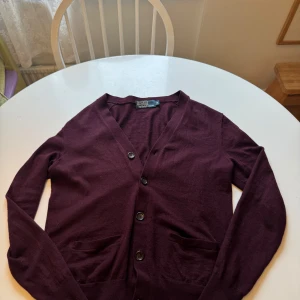 Vinröd kofta från Polo Ralph Lauren - Säljer en stilren vinröd kofta från Polo Ralph Lauren. Koftan har en v-ringning och knappar framtill. Perfekt för att hålla sig varm och snygg under kyligare dagar. Kom med bud! 