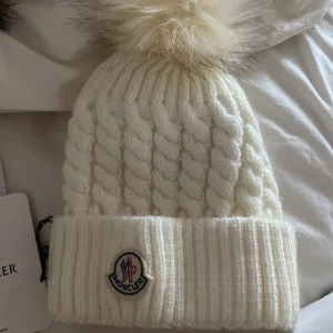 Vit stickad mössa från Moncler - Snygg vit stickad mössa från Moncler med en fluffig pälsboll på toppen. Mössan har ett klassiskt flätmönster och en uppvikt kant med Monclers logotyp. Perfekt för att hålla värmen med stil.