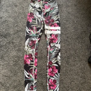Blommiga tights med tropiskt mönster - Snygga tränings tights med ett tropiskt blommönster i rosa, grönt och vitt på en svart bakgrund. Perfekta för att liva upp garderoben med lite färg och mönster. De har en tight passform och är perfekt för träning.