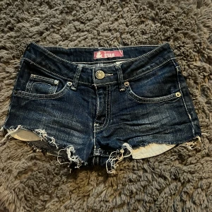 Mörkblå jeansshorts  - Säljer ett par mörkblå jeansshorts med fransiga kanter och klassisk femficksdesign. Perfekta för en avslappnad sommarlook. De har en knapp och dragkedja framtill.