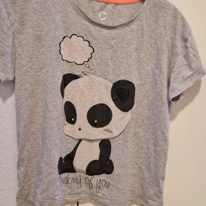 Grå t-shirt med panda-tryck - Säljer en söt grå t-shirt med ett stort panda-tryck och texten 'Thinking of you'. T-shirten har korta ärmar och en rund halsringning. Perfekt för en avslappnad stil! Nattplagg/T-shirt 