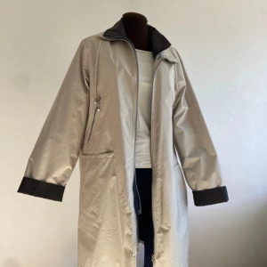 Beige kappa från Cappuccini - Snygg beige kappa från Cappuccini med dragkedja och långa ärmar. Jackan har en stilren design med mörka detaljer vid krage och ärmslut. Perfekt för en elegant look. Defekter enligt sista bild storlek är ca M-L (mer L)