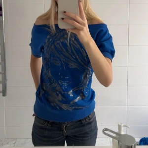 Blå T-shirt med tryck från Prime - Säljer en snygg blå offshoulder topp från Prime med ett tryck av ett ansikte i silver. Står ingen storlek men skulle säga S. Använd någon gång men jättebra skick