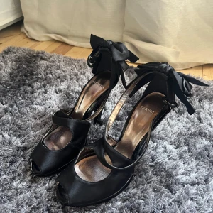 Svarta klackskor med rosett från Carvela - Eleganta svarta klackskor från Carvela med öppen tå och stora rosetter vid ankeln. Skorna har en stilren design med hög klack och är perfekta för en festlig kväll. Tillverkade i ett glansigt material som ger en lyxig känsla.
