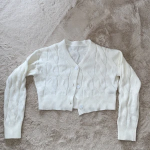 Dazy Vit stickad kofta - Säljer en söt vit stickad kofta med knappar framtill. Koftan är croppad och har ett diskret mönster med långa ärmar och ribbade muddar. Perfekt för lager-på-lager-stil! 🧶
