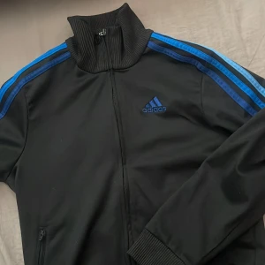 Svart träningsjacka från Adidas - Snygg svart träningsjacka från Adidas med blå ränder längs ärmarna och dragkedja framtill. Jackan har en hög krage och ribbade muddar vid ärmslut. Perfekt för sportiga aktiviteter eller en avslappnad stil. Nopprig passar XS men är egentligen storlek 152