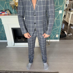 Grå rutig kostym från Digel - Helt ny kostym och Haupt skjorta ingår. Snygg grå rutig kostym från Digel med kavaj och matchande byxor. Kostymen har en klassisk passform med två knappar framtill och är perfekt för formella tillfällen. Materialet är en blandning av ull och polyester vilket ger en bekväm och stilren look.