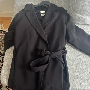 Svart kappa med bälte - Elegant svart kappa med bälte och knappar. Perfekt för en stilren look. Kappan har långa ärmar och en klassisk krage. Knapparna på insidan av kappan är in sydd med olika färger på trådarna men det är inget som syns utan på 
